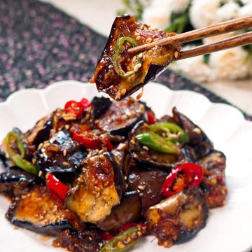 A plate of stir-fried Korean eggplant with gochujang, garnished with sliced red peppers and green onions, being picked up with chopsticks.