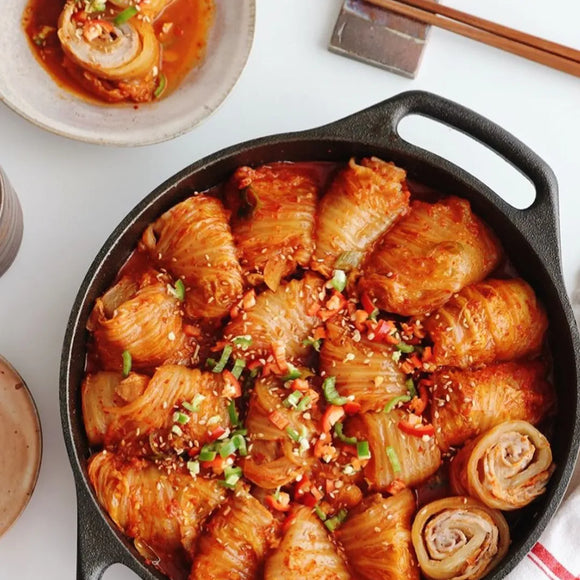 A pan of braised kimchi rolls with samgyup pork, garnished with sesame seeds and green onions, served alongside steamed rice and chopsticks.