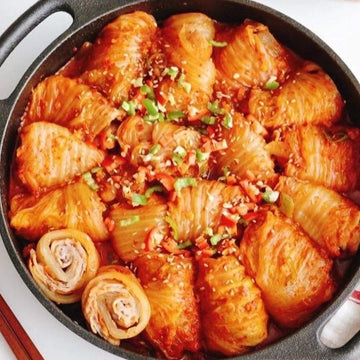 A platter of braised kimchi rolls with samgyup pork, garnished with green onions, sesame seeds, and red pepper flakes, served in a round black dish.