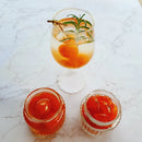 Glass of Kumquat Jelly cocktail with garnish and two jars of red jelly on a marble surface