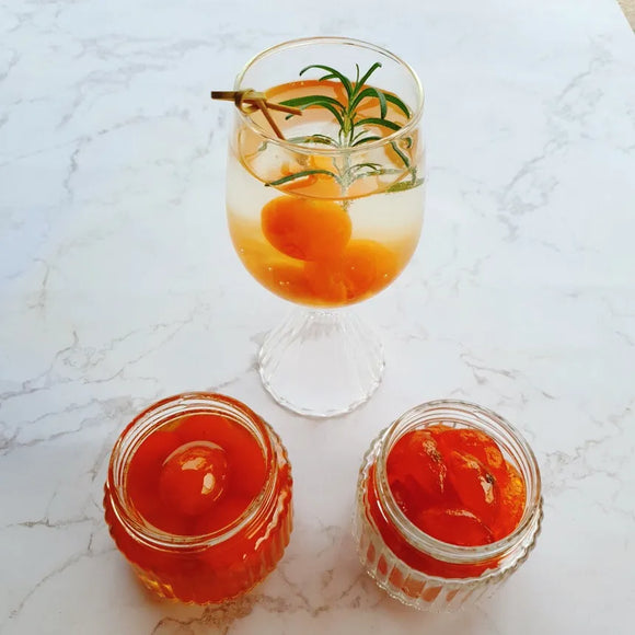 Glass of Kumquat Jelly cocktail with garnish and two jars of red jelly on a marble surface