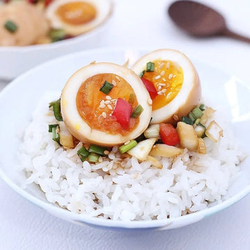 Bowl of rice with boiled eggs, vegetables, and sesame seeds on a white plate.