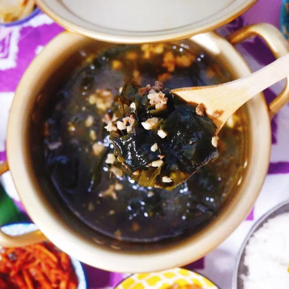 A bowl of Miyeokguk (Beef Seaweed Soup) with a wooden spoon lifting some seaweed and beef, garnished with sesame seeds, on a colorful tablecloth background.