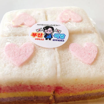Layered tteok with pink heart-shaped decorations and a visible brand label on a white background
