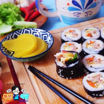 Pork Kimbap on a wooden board with a bowl of pickled radish and chopsticks, set against a colorful background.