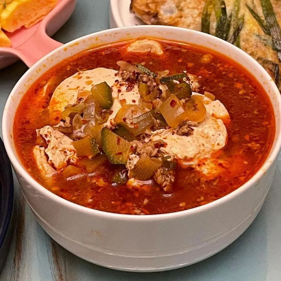 A bowl of Pork Sundubu Jjigae, featuring soft tofu, ground pork, egg, and scallions in a red broth.