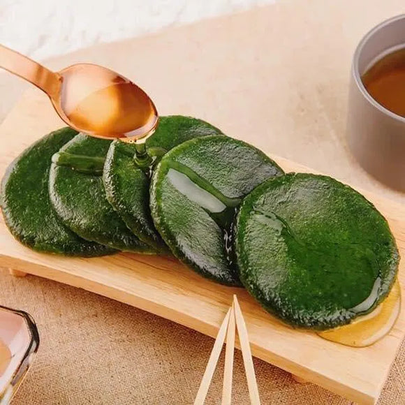Ssuk-gaeduk. Green rice cake on a wooden board with a spoon drizzling syrup.