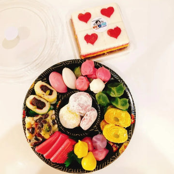 Assorted colorful tteoks on a black plate with a cake in the background