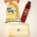 Packaged tteokbokki items including a bag of noodles, a bottle of sauce, and a package of sticks on a white background.