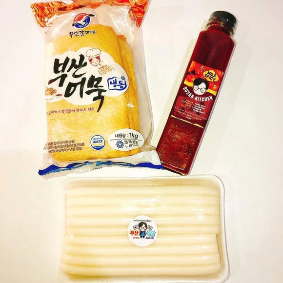 Packaged tteokbokki items including a bag of noodles, a bottle of sauce, and a package of sticks on a white background.
