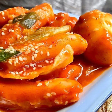 A plate of Tteokbokki, consisting of spicy rice cakes with gochujang sauce, fish cakes, and eggs, garnished with sesame seeds and scallions.